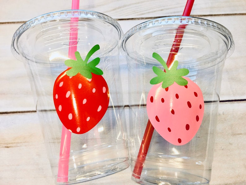 Puede incluir: Dos vasos de pl&aacute;stico transparentes con pajitas y dise&ntilde;os de fresas. Un vaso tiene una fresa roja y una pajita rosa, el otro tiene una fresa rosa y una pajita roja.