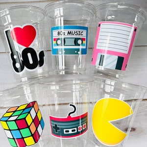 K&ouml;nnte beinhalten: Sechs durchsichtige Plastikbecher mit bunten Aufklebern zum Thema der 80er Jahre. Die Aufkleber zeigen ein Herz mit "I love 80s", ein Kassettenband mit "80s Music", eine Diskette, einen Rubik's Cube, einen Videospielcontroller und einen Pac-Man.