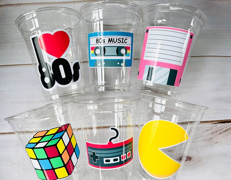 K&ouml;nnte beinhalten: Sechs durchsichtige Plastikbecher mit bunten Aufklebern zum Thema der 80er Jahre. Die Aufkleber zeigen ein Herz mit "I love 80s", ein Kassettenband mit "80s Music", eine Diskette, einen Rubik's Cube, einen Videospielcontroller und einen Pac-Man.