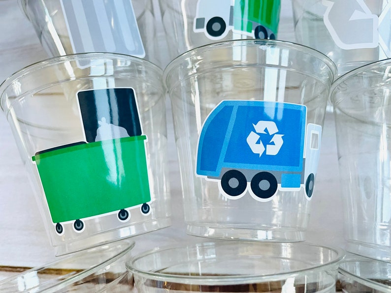 May include: Clear plastic cups with colorful stickers of garbage trucks and recycling bins. The stickers feature a blue recycling truck with a recycle symbol, a green garbage bin with a white bag, and a gray recycling bin with a white arrow.