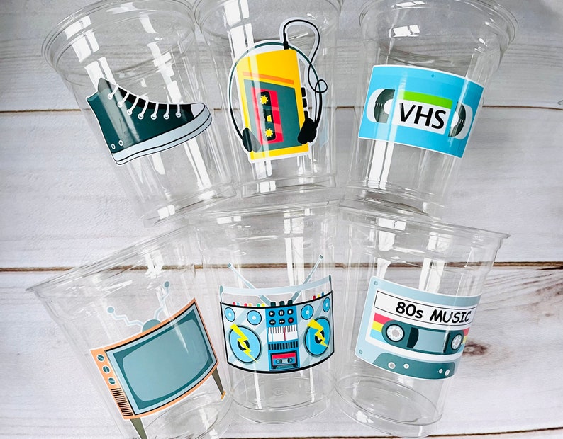 K&ouml;nnte beinhalten: Sechs durchsichtige Plastikbecher mit bunten Aufklebern im 80er-Jahre-Stil. Die Aufkleber zeigen ein Paar Turnschuhe, ein Kassettenband, eine Boombox, einen Fernseher und ein VHS-Band.