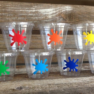 Peut inclure: Sept gobelets en plastique transparent avec des motifs de peinture &eacute;clabouss&eacute;s de couleurs. Les gobelets sont dispos&eacute;s en rang&eacute;e sur une surface en bois. Les couleurs sont le rouge, l'orange, le jaune, le vert, le bleu et le violet.