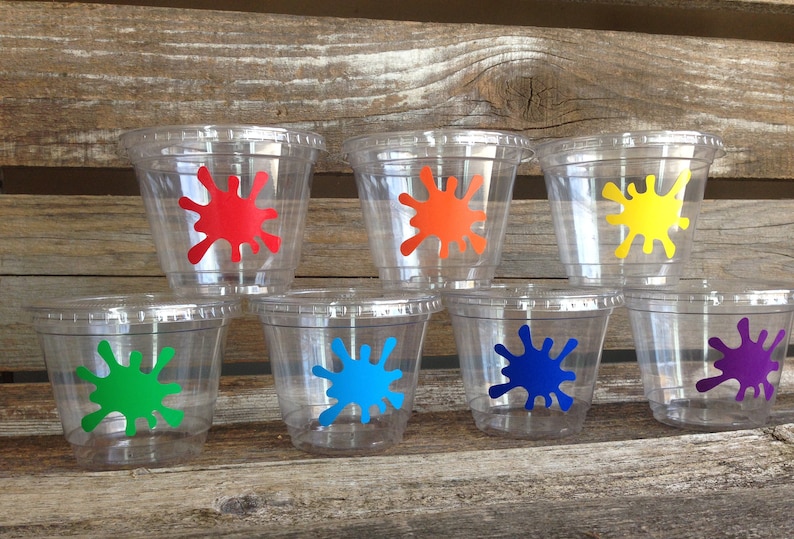 Peut inclure: Sept gobelets en plastique transparent avec des motifs de peinture &eacute;clabouss&eacute;s de couleurs. Les gobelets sont dispos&eacute;s en rang&eacute;e sur une surface en bois. Les couleurs sont le rouge, l'orange, le jaune, le vert, le bleu et le violet.