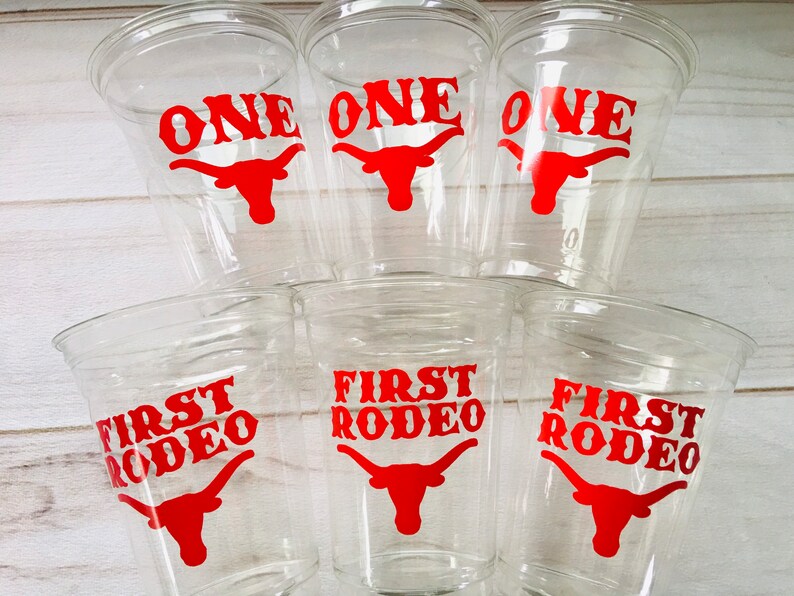 K&ouml;nnte beinhalten: Sechs durchsichtige Plastikbecher mit roten und wei&szlig;en Designs. Drei Becher sagen "ONE" mit einem roten Longhorn-Stierkopf. Drei Becher sagen "FIRST RODEO" mit einem roten Longhorn-Stierkopf.