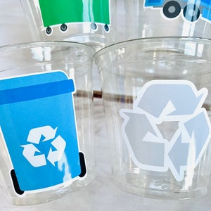 May include: Four clear plastic cups with recycling themed stickers. One cup has a blue recycling bin with a white recycling symbol. Another cup has a gray recycling symbol. The other two cups have stickers that are not visible in the image.