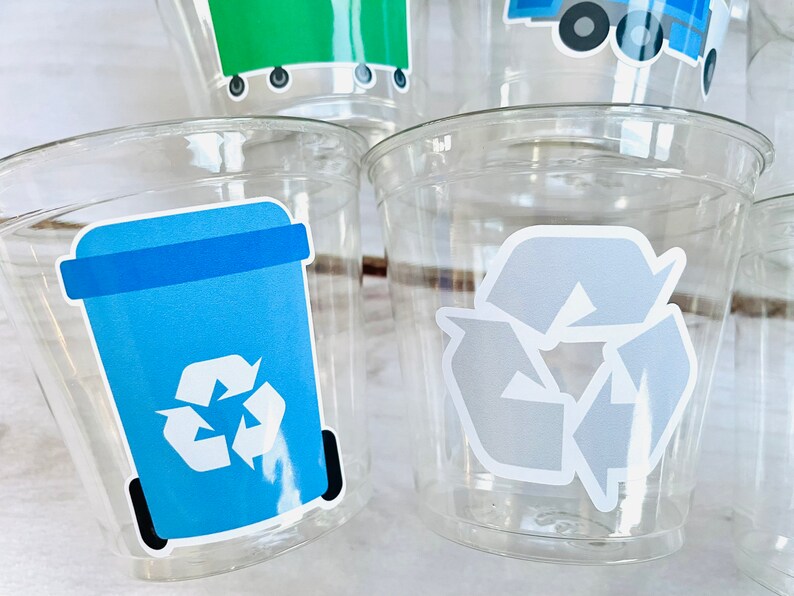 May include: Four clear plastic cups with recycling themed stickers. One cup has a blue recycling bin with a white recycling symbol. Another cup has a gray recycling symbol. The other two cups have stickers that are not visible in the image.