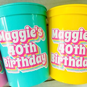 Puede incluir: Primer plano de tres vasos de pl&aacute;stico en rosa, turquesa y amarillo. Cada vaso tiene una pegatina con la frase "Maggie's 40th Birthday" en una fuente retro colorida. Adecuado para una fiesta.