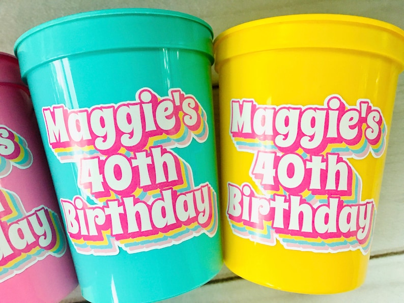 Puede incluir: Primer plano de tres vasos de pl&aacute;stico en rosa, turquesa y amarillo. Cada vaso tiene una pegatina con la frase "Maggie's 40th Birthday" en una fuente retro colorida. Adecuado para una fiesta.