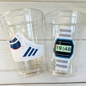 K&ouml;nnte beinhalten: Zwei durchsichtige Plastikbecher mit wei&szlig;en und blauen Aufklebern. Ein Becher hat einen Aufkleber mit einem wei&szlig;en Sneaker mit blauen und roten Akzenten. Der andere Becher hat einen Aufkleber mit einer Digitaluhr mit einem grau-blauen gestreiften Band und einem gr&uuml;nen Digitaldisplay, das "19:48" anzeigt.