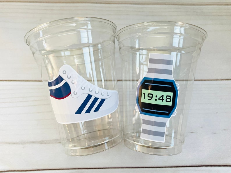 K&ouml;nnte beinhalten: Zwei durchsichtige Plastikbecher mit wei&szlig;en und blauen Aufklebern. Ein Becher hat einen Aufkleber mit einem wei&szlig;en Sneaker mit blauen und roten Akzenten. Der andere Becher hat einen Aufkleber mit einer Digitaluhr mit einem grau-blauen gestreiften Band und einem gr&uuml;nen Digitaldisplay, das "19:48" anzeigt.