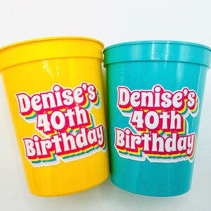 Puede incluir: Dos vasos de pl&aacute;stico, uno amarillo y otro turquesa, con el texto "Denise's 40th Birthday" en una fuente colorida de estilo retro. Los vasos son probablemente para una fiesta de cumplea&ntilde;os.