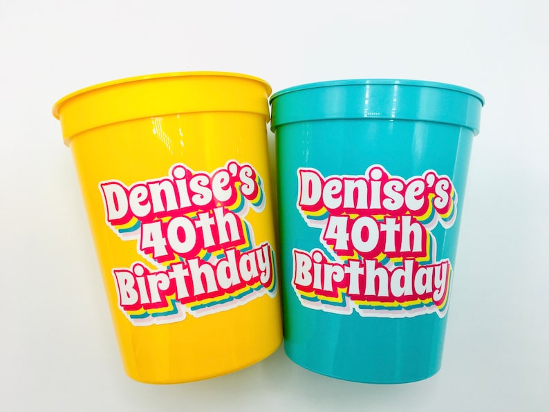 Puede incluir: Dos vasos de pl&aacute;stico, uno amarillo y otro turquesa, con el texto "Denise's 40th Birthday" en una fuente colorida de estilo retro. Los vasos son probablemente para una fiesta de cumplea&ntilde;os.