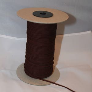 Peut inclure: Une bobine de cordon brun foncé, enroulée autour d'un noyau en carton. Le cordon est de couleur unie, et une longueur s'étend de la bobine. La bobine est posée sur une surface blanche.