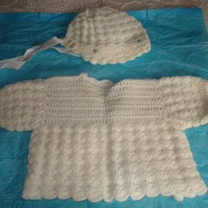 May include: A white crocheted baby sweater and hat set. The sweater has a scalloped edge and the hat has a small white button.