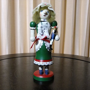 May include: A wooden nutcracker doll with a green dress, white lace collar, and red and white trim. The doll is holding a smaller wooden nutcracker doll in its right hand. The doll is standing on a round wooden base.