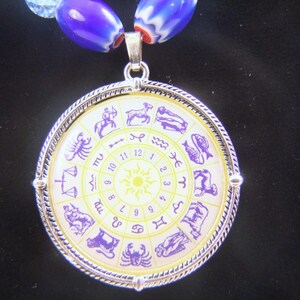 May include: A silver pendant necklace with a round, white and yellow zodiac wheel charm. The charm features purple zodiac symbols and numbers 1 through 12 around the edge.