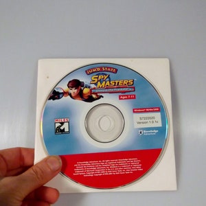 May include: A CD for the computer game "JumpStart Spy Masters: Unmask the Phantom" by Knowledge Adventure. The CD is white with a red border and features a cartoon character in a blue jumpsuit. The game is for ages 7-11. The CD is labeled "Windows 98/Me/2000" and "S7222020 Version 1.0.1c".
