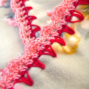 May include: Close-up of a handmade crochet trim in shades of pink and red. The trim features a scalloped edge and is set against a soft, floral patterned background. This decorative trim is suitable for various craft projects.