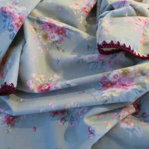 May include: Pale green fabric with a floral design in pink, yellow, and white. The edges are trimmed with a dark red crocheted border. The fabric is folded, showing the pattern of flowers. Suitable for home textiles.