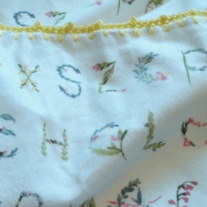 May include: White fabric with floral letter designs in green, pink, and blue. The letters are decorated with leaves and flowers. A yellow crocheted trim borders the top edge of the fabric.