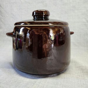 May include: A dark brown ceramic pot with a matching lid. The pot has two small handles on either side and a rounded shape. The lid has a small knob on top for easy removal. The pot is likely used for cooking or storage.
