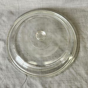 May include: A clear glass lid with a small knob in the center. The lid is round with a slightly raised edge and is sitting on a gray fabric surface. The lid appears to be for a pot or pan.