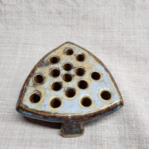 May include: A ceramic flower frog with a triangular shape and multiple holes for arranging stems. The frog has a light blue glaze with brown and tan accents. The base is a darker brown color.
