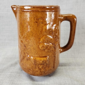 May include: A brown ceramic pitcher with a handle and a spout. The pitcher has a textured surface and embossed designs. The overall shape is cylindrical with a rounded base. The pitcher is likely vintage or antique.
