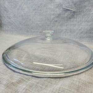 May include: A clear glass lid with a small, decorative handle. The lid is circular and appears to be for a pot or pan. The glass is transparent, allowing a view of the contents below. The handle is a small, clear, faceted knob.