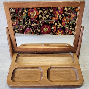 May include: A vintage wooden vanity mirror with a rectangular frame and a decorative floral fabric backing. The mirror has a small tray with three compartments for storing jewelry or small items. The wood has a warm, natural finish.