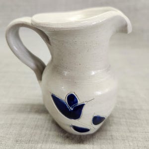 May include: A small, off-white ceramic pitcher with a curved handle and spout. The pitcher features a blue floral design on the front. The handmade pottery has a rustic, speckled texture, suggesting a handcrafted origin.