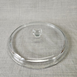May include: A clear glass lid with a small, square handle. The lid is circular with a raised rim and is designed to fit on a pot or pan. The glass is transparent, allowing visibility of the contents.
