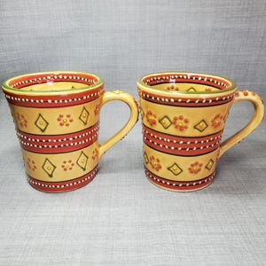 May include: Two ceramic mugs with a yellow base and red, green, and yellow geometric designs. The mugs have a white interior and a rounded handle.