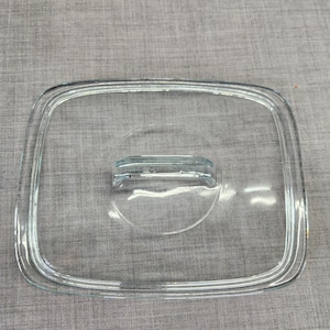 May include: A rectangular, clear glass lid featuring rounded corners and a centered, integrated glass handle. The lid has a raised, stepped rim design around the perimeter, providing a secure fit for a corresponding container or cookware piece.