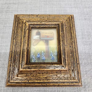 May include: A framed miniature painting depicts a mailbox with the name "RICHARDSON" on it, set against a backdrop of bluebonnets. The frame is made of wood with a rustic, textured finish, adding a touch of country charm to the artwork.