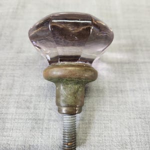 May include: A vintage door knob with a faceted, light purple glass head. The knob sits atop a tarnished brass base and a threaded metal rod. The hardware is likely from an antique door or cabinet.