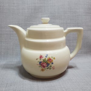 May include: A white ceramic teapot with a floral design. The teapot has a lid and a handle. The floral design features pink, yellow, and blue flowers.