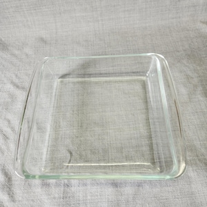 May include: A clear, square glass baking dish with rounded corners. The dish is empty and sits on a gray surface. The dish is designed for baking and cooking.
