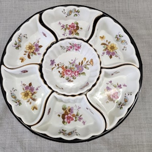 May include: A white, floral-patterned serving platter with a black rim. The platter is divided into seven sections, with a central round bowl and six surrounding petal-shaped compartments. Each section features a delicate floral design in shades of pink, yellow, and purple.