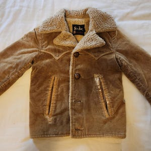May include: A tan corduroy jacket with a faux fur collar and lining. The jacket has two front pockets, a button closure, and decorative stitching. The label inside the jacket reads "Sir Jac".