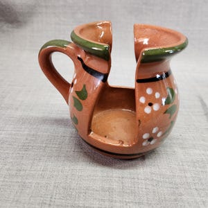 May include: A terracotta-colored ceramic candle holder with a handle and a split top. The holder is decorated with white floral patterns and green accents. Black lines outline the top and base.