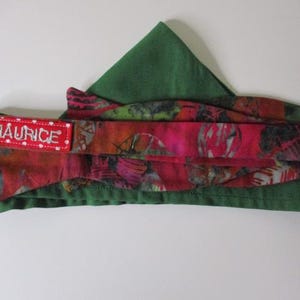 May include: A bow tie featuring a vibrant, multi-colored abstract pattern in shades of pink, red, and black, layered over a solid dark green fabric. A rectangular red fabric tag with white polka dots and the text JMAURICE is attached to the side.