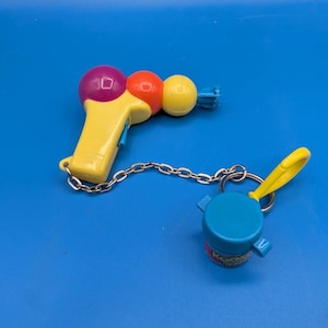 May include: A yellow plastic toy blaster with a purple and orange sphere on top, connected by a silver metal chain to a blue cylindrical container with a yellow plastic clip. The container features the text Koosh printed on its side.