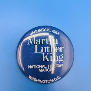 May include: A round, blue button with white text. The button reads "January 15, 1982" at the top, with "Martin Luther King" in the center, and "NATIONAL HOLIDAY MARCH WASHINGTON, D.C." at the bottom. The button is set against a solid blue background.