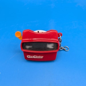 May include: A red plastic View-Master keychain featuring a yellow advance lever on the left side and a metal split ring attached to the right. The front of the device displays the white View-Master logo on a red background with two black viewing lenses.