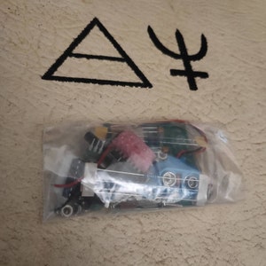 May include: A clear plastic bag containing electronic components, including circuit boards, wires, and other small parts. The background features a beige surface with black geometric symbols.