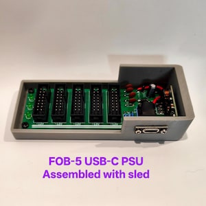 Peut inclure: Une carte de circuit imprimé verte avec cinq connecteurs noirs et un port USB-C montés dans un boîtier en plastique gris imprimé en 3D. La carte comporte des composants électroniques, dont des condensateurs et des résistances. Le texte sur la carte indique -12V. Le texte en dessous indique FOB-5 USB-C PSU Assembled with sled.