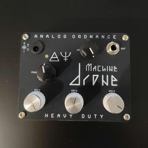 Puede incluir: Pedal de efectos negro "MACHINE DRONE" con perillas plateadas y letras blancas. El pedal tiene el texto "ANALOG ORDNANCE" y "HEAVY DUTY". Cuenta con múltiples conectores de entrada y salida y perillas de control.