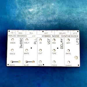 May include: A white electronic circuit board with black text and various ports and knobs. The text includes "ANALOG ORDNANCE," "HYBRID NOISE SYNTHESIZER," and labels like "OSC," "VOLT," and "RATE." The board is on a blue surface.