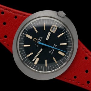May include: A silver Omega Geneve Dynamic automatic wristwatch with a black face and a red leather band with holes. The watch face has gold-colored hour markers and a blue second hand.